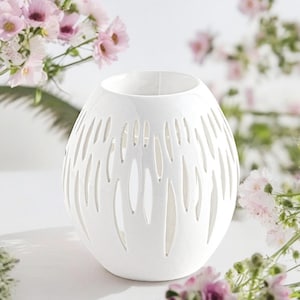 Peut inclure: Un bougeoir en céramique blanche de forme arrondie, rappelant un œuf, doté de découpes verticales allongées sur tout le corps. La surface est lisse et mate, avec une large ouverture circulaire au sommet pour une bougie ou une source lumineuse.