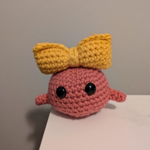 Op de afbeelding: Een gehaakt pluche speelgoed met een roze lichaam en een gele strik. Het speelgoed heeft zwarte knoopjes voor ogen.