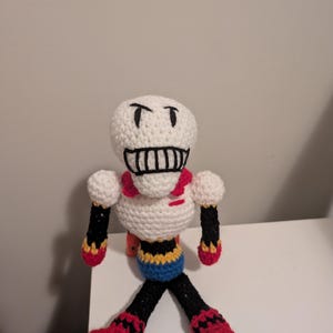 May include: A handmade crochet doll of a skeleton character. The doll has a white head with black eyes and a mouth, a red scarf, and a black and yellow torso. The legs and arms are black with red accents. The doll is approximately 10 inches tall.