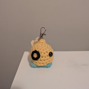 Puede incluir: Un llavero de pescado de crochet amarillo y azul con ojos negros. El pez tiene una cola pequeña y un llavero plateado adjunto.