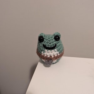 Puede incluir: Un pequeño juguete de rana de crochet con ojos negros y una barriga a rayas marrones y blancas.