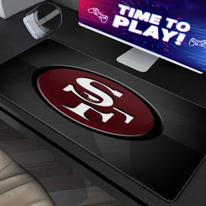 May include: A rectangular black desk mat featuring a large, centered oval emblem with a dark red textured background and the white San Francisco 49ers logo. The mat has stitched edges and is positioned on a desk surface below a computer monitor.
