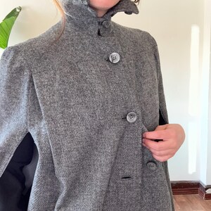 Puede incluir: Una capa de lana gris con cuello alto y cierre de botones. La capa tiene una silueta larga y fluida y es perfecta para superponer sobre un vestido o un su&eacute;ter.