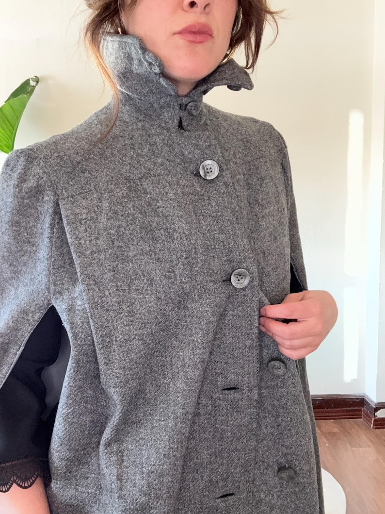 Puede incluir: Una capa de lana gris con cuello alto y cierre de botones. La capa tiene una silueta larga y fluida y es perfecta para superponer sobre un vestido o un su&eacute;ter.