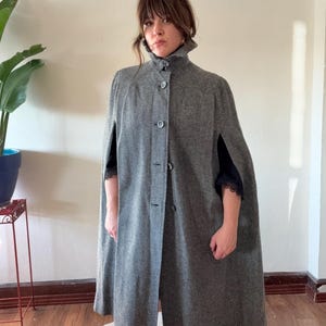 Puede incluir: Una capa de lana gris con cuello alto y cierre de botones. La capa tiene una silueta larga y fluida y es perfecta para superponer sobre un vestido o un su&eacute;ter.