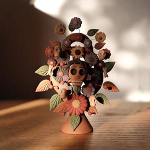 Może przedstawiać: Ręcznie wykonana rzeźba z gliny przedstawiająca centralną postać z twarzą czaszki, otoczoną kolorowymi kwiatami i małymi czaszkami. Rzeźba ma terakotową podstawę i jest ozdobiona zielonymi, czerwonymi i fioletowymi elementami kwiatowymi.