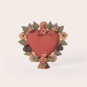 May include: A terracotta-colored heart-shaped sculpture, adorned with a wreath of clay flowers and leaves in shades of green, orange, and purple. The sculpture is mounted on a small, cone-shaped base. The overall design is whimsical and decorative.