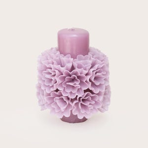 Puede incluir: Una vela de color lavanda con un diseño floral. La vela tiene forma cilíndrica con un pilar central y está rodeada de capas de pétalos de flores esculpidos. La estética general es suave y decorativa, adecuada para la decoración del hogar.