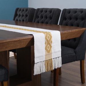 Peut inclure: Chemin de table blanc et doré avec un motif géométrique et des franges. Le chemin de table est posé sur une table en bois, avec des chaises rembourrées gris foncé en arrière-plan. Le motif du chemin de table présente un motif répétitif doré.