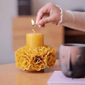 Puede incluir: Una vela amarilla encendida se encuentra sobre un portavelas floral. Se utiliza una cerilla para encender la vela. En el fondo, hay una taza de cerámica decorativa con un diseño de rostro. El portavelas y la vela están sobre una superficie de madera.