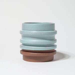 May include: A two-toned ceramic vase with a unique, ridged design. The top portion is a soft, muted teal color, while the base is a warm, earthy brown. The vase has a cylindrical shape and is suitable for displaying flowers or as a decorative piece.
