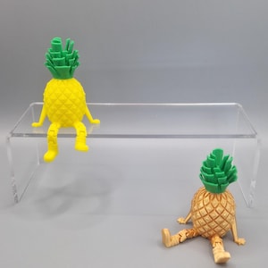 Könnte beinhalten: Zwei Ananas-Figuren mit grünen Oberteilen und gelben Körpern, die auf einem durchsichtigen Acrylregal sitzen. Eine Ananas sitzt aufrecht mit herabhängenden Beinen, während die andere mit ausgebreiteten Beinen liegt.