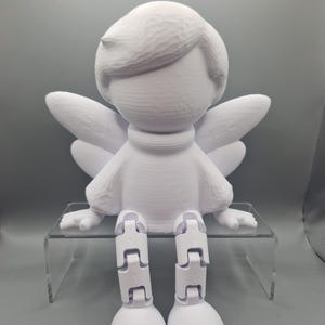 Könnte beinhalten: Eine weiße, 3D-gedruckte Engelsfigur mit beweglichen Beinen und Flügeln. Der Engel hat ein stilisiertes Gesicht, kurzes Haar und trägt eine Tunika. Die Figur sitzt auf einem transparenten Acrylregal vor grauem Hintergrund.