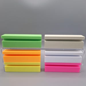 Könnte beinhalten: Eine Sammlung farbenfroher, rechteckiger Seifenschalen in Grün, Orange, Gelb, Beige, Weiß und Pink. Jede Schale hat ein geschlitztes Design zur Entwässerung. Die Seifenschalen sind paarweise gestapelt und zeigen eine Vielzahl lebendiger Farben.