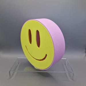 Könnte beinhalten: Eine dreidimensionale Smiley-Gesichts-Skulptur. Das Gesicht ist gelb mit einem rosa Rand und Seiten. Die Augen und der Mund sind ausgeschnitten. Die Skulptur wird auf einem transparenten Acrylständer ausgestellt.