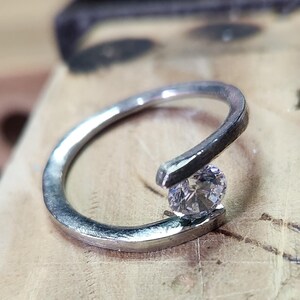 May include: A silver ring with a single, round, clear gemstone set in the band. The ring is designed with a curved band.