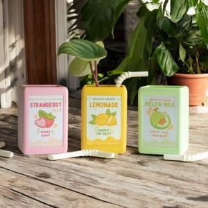 Op de afbeelding: Drie kleurrijke, cartoonachtige sapdozen met rietjes. De dozen zijn roze, geel en groen en bevatten illustraties van aardbeien, citroenen en meloenen. De tekst op de dozen luidt "Strawberry Milk", "Lemonade" en "Melon Milk".