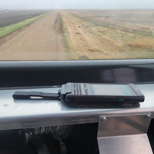 May include: A black handheld scanner with a keypad and a screen, sitting on a silver surface. The scanner is in focus, while the background is blurry and shows a dirt road leading into a field.