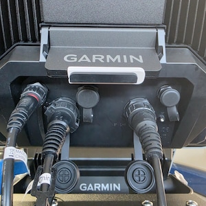 May include: A close-up shot of a black Garmin marine electronics unit. The device features multiple ports and connectors, with the Garmin logo visible. Cables are connected to the unit. The device is mounted on a black stand.