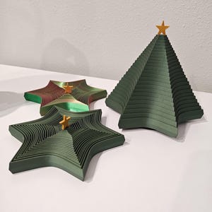 May include: Three green paper Christmas trees, two are star-shaped and one is a traditional pyramid shape. Each tree is made of multiple layers of paper and has a gold star on top.