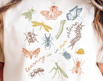 May include: Ivory-colored t-shirt featuring a collection of illustrated insects in various colors. The shirt includes a dragonfly, butterfly, and ant, with the text "learning is in our nature" in a handwritten style. The shirt is made of a soft material.