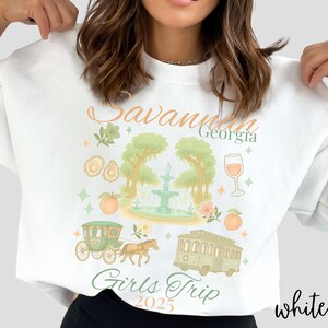 May include: White sweatshirt with a colorful graphic design featuring the text "Savannah Georgia Girls Trip 2025." The design includes illustrations of a fountain, trees, a horse-drawn carriage, a trolley, and peaches.