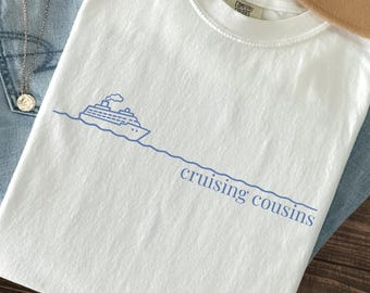 May include: White t-shirt with a blue graphic of a cruise ship and the words "cruising cousins." The shirt is displayed on a wooden surface with blue jeans and a necklace.