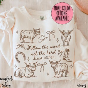 May include: Ivory-colored sweatshirt with a brown graphic design featuring Highland cows, a cowboy hat, bows, a horseshoe, and a cowboy boot. Text reads "Follow the word not the herd Isaiah 8:11-13." The sweatshirt has a crew neck and long sleeves.