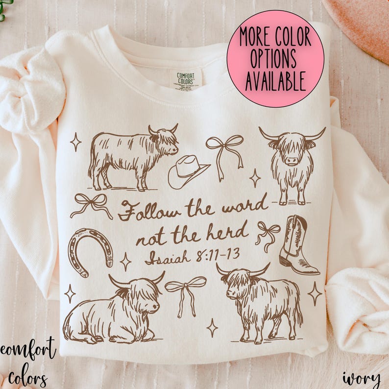 May include: Ivory-colored sweatshirt with a brown graphic design featuring Highland cows, a cowboy hat, bows, a horseshoe, and a cowboy boot. Text reads "Follow the word not the herd Isaiah 8:11-13." The sweatshirt has a crew neck and long sleeves.
