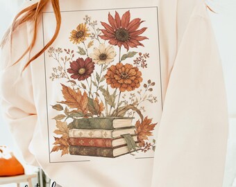 Puede incluir: Una sudadera de color marfil con un gráfico rectangular que muestra una pila de cuatro libros coronada por un ramo de flores en tonos otoñales y hojas secas. Las flores incluyen grandes girasoles de color óxido, zinnias naranjas y pequeñas flores blancas.