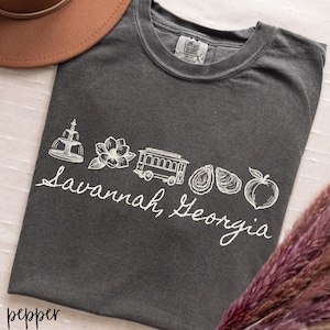 Puede incluir: Una camiseta gris oscuro con un dise&ntilde;o gr&aacute;fico blanco que presenta una fuente, una flor, un tranv&iacute;a, ostras y un melocot&oacute;n, con el texto "Savannah, Georgia" en cursiva. La camiseta est&aacute; etiquetada como "pepper".