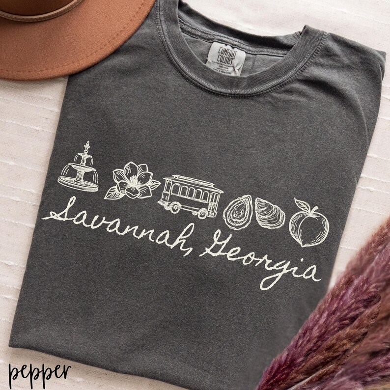 Puede incluir: Una camiseta gris oscuro con un dise&ntilde;o gr&aacute;fico blanco que presenta una fuente, una flor, un tranv&iacute;a, ostras y un melocot&oacute;n, con el texto "Savannah, Georgia" en cursiva. La camiseta est&aacute; etiquetada como "pepper".