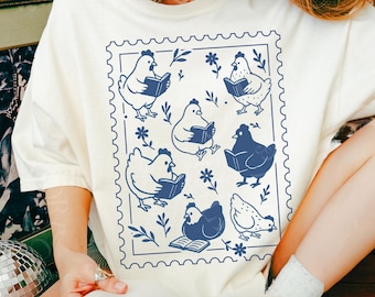 May include: Ivory-colored t-shirt featuring a blue stamp design with cartoon chickens reading books. The design includes floral accents and a scalloped border. The shirt is a casual, comfortable style.