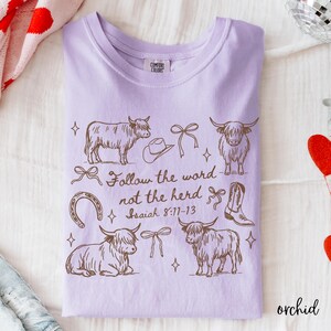 May include: Orchid-colored t-shirt with a brown graphic design featuring Highland cows, a cowboy hat, bows, a horseshoe, and a cowboy boot. The text "Follow the word not the herd" is also printed on the shirt.