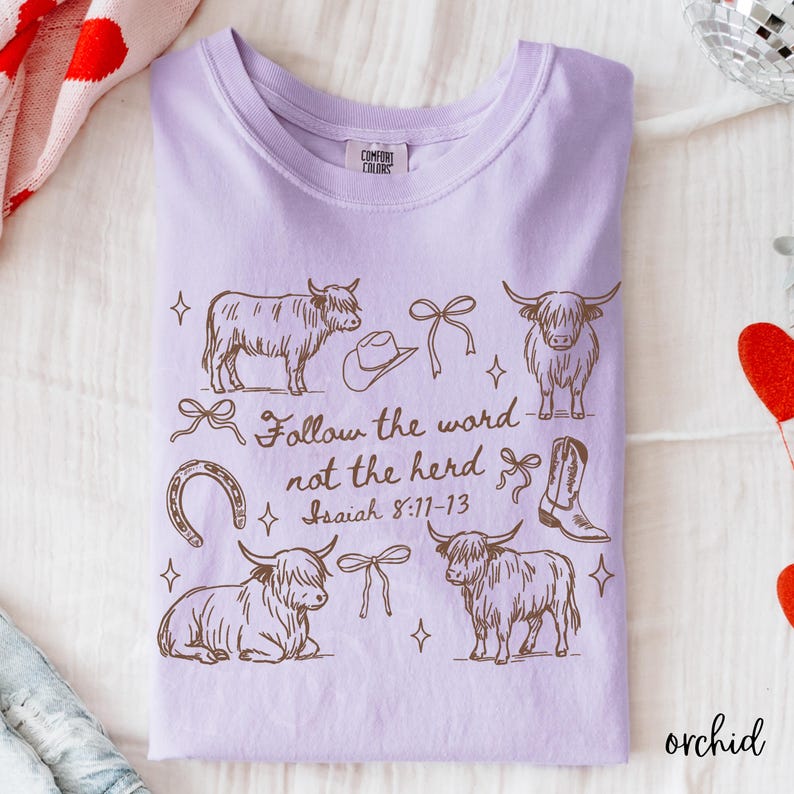 May include: Orchid-colored t-shirt with a brown graphic design featuring Highland cows, a cowboy hat, bows, a horseshoe, and a cowboy boot. The text "Follow the word not the herd" is also printed on the shirt.