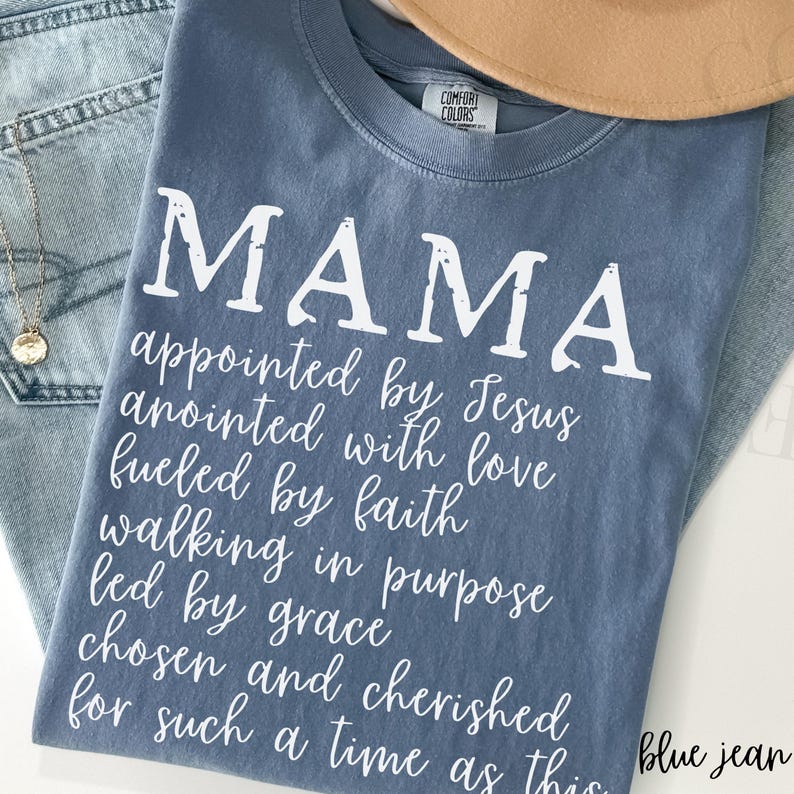 May include: A blue Comfort Colors t-shirt with the word "MAMA" in large white letters. Below, a list of inspirational words is printed in white script. The shirt is folded on top of blue jeans.