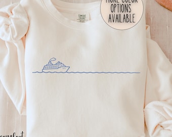 May include: Ivory-colored sweatshirt with a blue line drawing of a cruise ship on the ocean. The text "MORE COLOR OPTIONS AVAILABLE" is in a circle at the top right. The brand tag says "COMFORT COLORS".