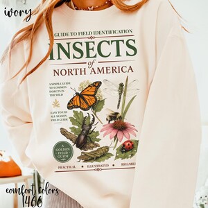 May include: An ivory sweatshirt featuring a graphic print titled Guide to Field Identification Insects of North America. The design includes detailed illustrations of a monarch butterfly, dragonfly, beetle, bee, grasshopper, and ladybug on a flower.