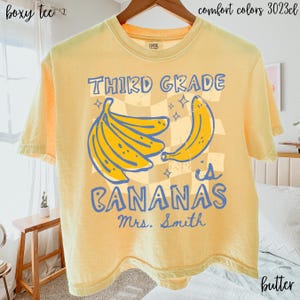 May include: A pale yellow boxy t-shirt featuring a graphic print with the text "THIRD GRADE is BANANAS" and "Mrs. Smith". The design includes a yellow banana bunch and a single banana over a light tan checkered background with small star accents.