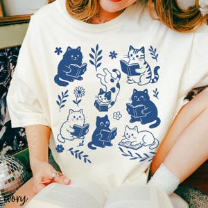 以下が含まれることがあります： クリーム色のTシャツ。本を読む猫のネイビーブルーのパターンが繰り返され、花柄や葉のモチーフが散りばめられています。猫は様々なポーズで描かれており、デザインに視覚的な面白さを加えています。カジュアルなスタイルです。