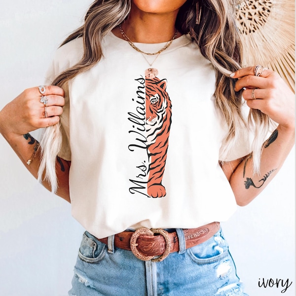 May include: A white t-shirt with a tiger graphic and the text "Mrs. Williams" printed on the front. The shirt is being worn by a person with blonde hair and is paired with blue jeans and a brown belt.