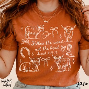 May include: Burnt orange t-shirt with a white graphic design featuring Highland cows, a cowboy hat, a horseshoe, and a cowboy boot. The text "Follow the word not the herd" is also included. The shirt is part of the "comfort colors" collection.