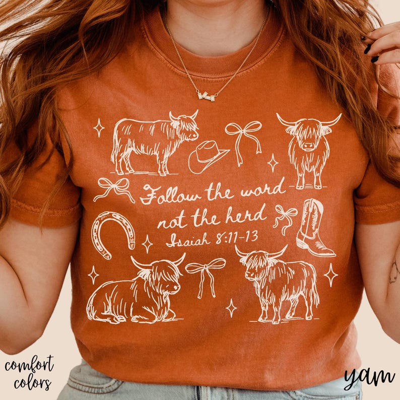 May include: Burnt orange t-shirt with a white graphic design featuring Highland cows, a cowboy hat, a horseshoe, and a cowboy boot. The text "Follow the word not the herd" is also included. The shirt is part of the "comfort colors" collection.