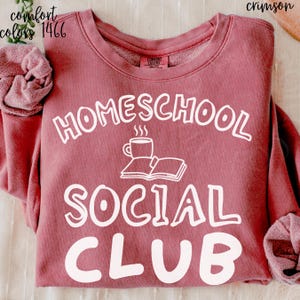 Op de afbeelding: Een karmozijnrood sweatshirt met ronde hals en witte opdruk HOMESCHOOL SOCIAL CLUB. In het midden staat een witte lijntekening van een open boek met een dampende kop koffie of thee erop.