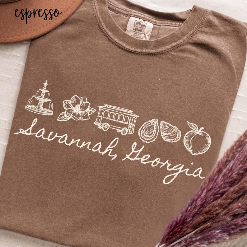 Puede incluir: Camiseta marr&oacute;n con un dise&ntilde;o blanco de Savannah, Georgia, que incluye una fuente, una flor, un tranv&iacute;a, ostras y un melocot&oacute;n. Tambi&eacute;n incluye el texto "Savannah, Georgia".