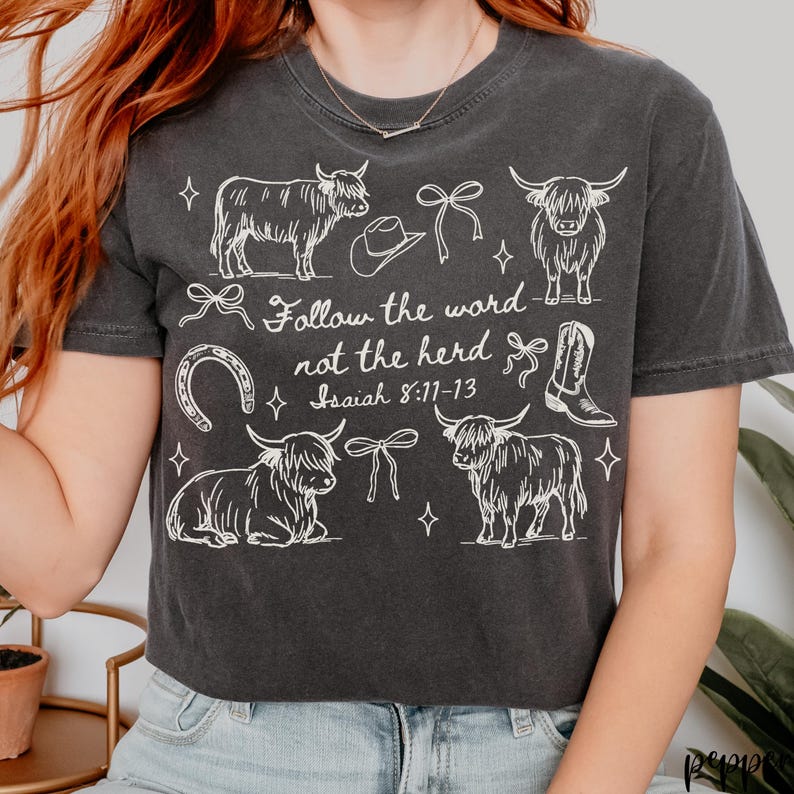May include: Dark gray t-shirt with a white graphic design featuring Highland cows, a cowboy hat, a horseshoe, and a cowboy boot. The text "Follow the word not the herd Isaiah 8:11-13" is also printed on the shirt.