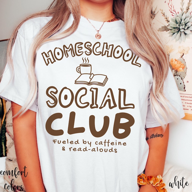 Op de afbeelding: Een wit T-shirt met korte mouwen en bruine tekst HOMESCHOOL SOCIAL CLUB, inclusief een afbeelding van een koffiemok en een open boek. Daaronder staat de kleinere bruine tekst Fueled by caffeine & read-alouds.