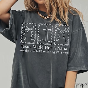 Pu&ograve; includere: T-shirt grigio scuro con la scritta "Jesus Made Her A Nana and she wouldn't have it any other way." La maglietta presenta tre disegni bianchi incorniciati: due fiocchi e una croce.