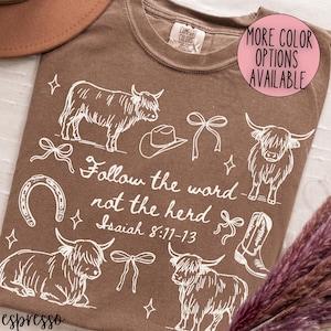 May include: Espresso-colored t-shirt with a white graphic design featuring Highland cows, a cowboy hat, bows, a horseshoe, and a cowboy boot. The text "Follow the word not the herd Isaiah 8:11-13" is also included. A pink circle indicates more color options.