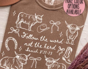 May include: A brown t-shirt with a white graphic of Highland cows, a cowboy hat, bows, a horseshoe, and a cowboy boot. The text "Follow the word not the herd Isaiah 8:11-13" is included. A pink circle indicates more colour options.